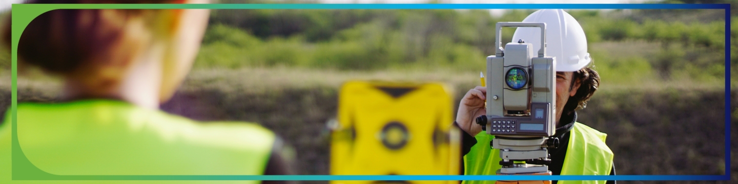 Two workers wearing high-visibility safety vests and hard hats are conducting a land survey outdoors. One person is operating a total station mounted on a tripod, while the other holds a survey prism target. The background shows grassy terrain and a slightly elevated area under natural daylight.