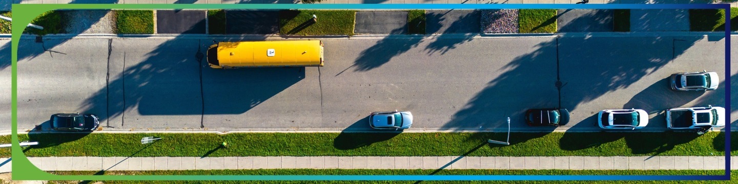 "Aerial view of a street with a yellow school bus and several parked cars, bordered by green grass and sidewalks, with shadows indicating a sunny day.
