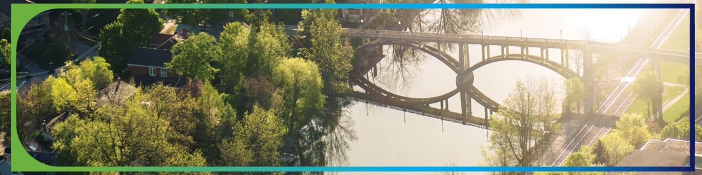 The image shows an aerial view of a river with a bridge crossing over it. The bridge has an arch design and casts a reflection on the water below. Surrounding the river are trees with green foliage, indicating a lush environment. There are also buildings visible on both sides of the river, suggesting an urban or suburban setting. The sunlight is bright, creating reflections and shadows in the scene.