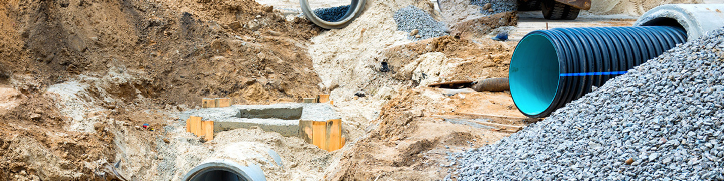 A construction site with large underground pipes being installed. The area has excavated sections, piles of dirt and gravel, and features a prominent blue pipe on the right alongside several concrete pipes.