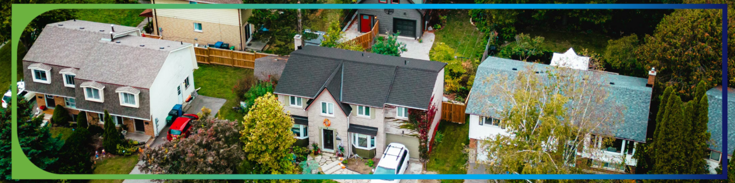Aerial view of a residential neighborhood showing three houses surrounded by trees and greenery. The central house has a black roof, while the other two have lighter-colored roofs. Driveways and parked cars are visible in front of the houses.