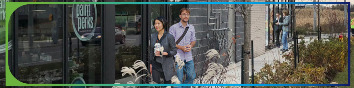 A street scene showing two people walking on a sidewalk in front of a building with large windows and a dark exterior. In the background, two other people are standing and talking near the entrance of another building.