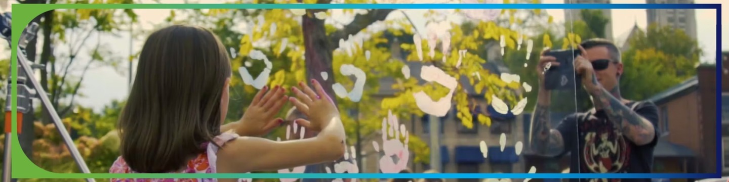 A young girl in a pink dress presses her hands against a glass surface with white handprints painted on it, while an adult with tattooed arms photographs her with a smartphone. Trees and buildings are visible in the background.
