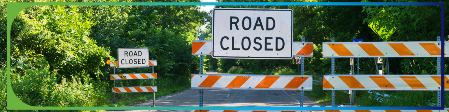 A road is blocked by multiple barricades with orange and white diagonal stripes. Two signs read 'ROAD CLOSED,' one in the foreground and one further back. The road is surrounded by dense green foliage on both sides.