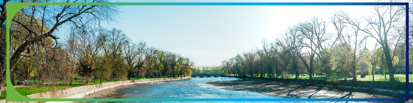 A wide river flowing gently under a bright blue sky, bordered by leafless trees and green grass on both sides, with sunlight reflecting off the water’s surface.