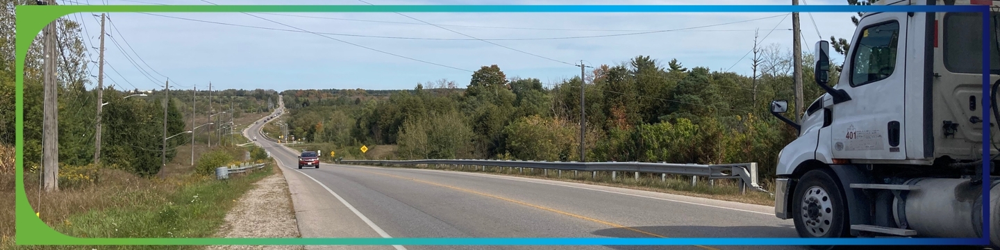 A rural road with a slight curve, bordered by utility poles and dense foliage. A red car approaches in the left lane while a white truck moves away on the right. The sky is mostly clear with light clouds.