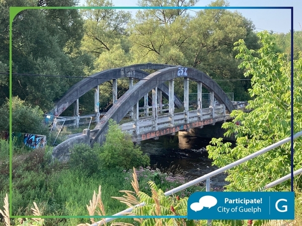 An old arched concrete bridge with graffiti, spanning a small river and surrounded by lush greenery. A chain-link fence is partially visible on the left.