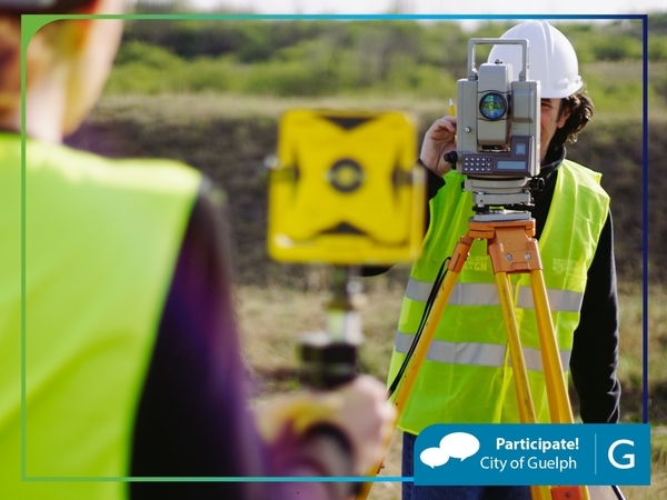 Two workers wearing high-visibility safety vests and hard hats are conducting a land survey outdoors. One person is operating a total station mounted on a tripod, while the other holds a survey prism target. The background shows grassy terrain and a slightly elevated area under natural daylight.
